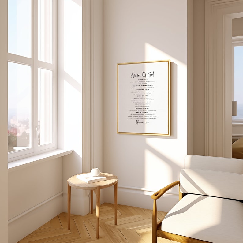 May include: A white framed print with the text "Armor of God" and a list of spiritual armour. The print is hanging on a white wall in a room with a wooden floor, a white armchair, and a small wooden table with a book and a white object on top.