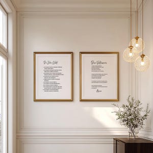 May include: Two framed art prints with gold frames hang on a white wall. The prints feature black text on a white background. The text appears to be a poem or a list. A decorative light fixture and a vase of flowers are also visible.