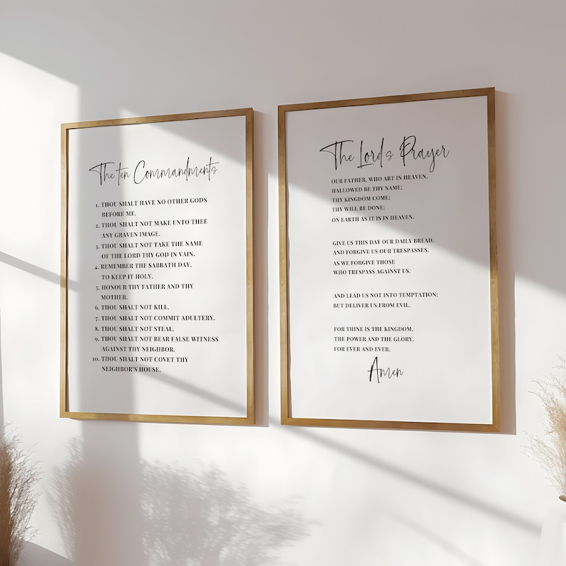 May include: Two framed art prints with gold-colored frames. The left print displays "The Ten Commandments" and the right print displays "The Lord's Prayer", both in elegant black script on a white background. The prints are hung on a white wall.