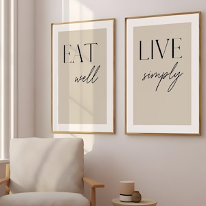以下が含まれることがあります： 「EAT well」と「LIVE simply」というフレーズが黒い筆記体で書かれた、額入りのウォールアートプリント2枚。プリントはベージュの背景で、金色のフレームに収められています。このアートワークは、ニュートラルカラーの壁に飾られています。