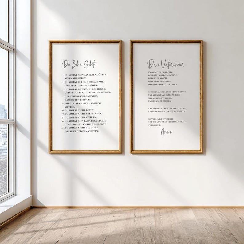 May include: Two framed prints with gold-colored frames hang on a white wall. The prints feature black text on a white background. The left print is titled "Du Zahn Gebote" and the right print is titled "Das Vaterunser". The prints are displayed near a window.
