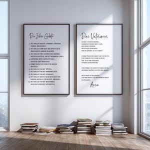May include: Two framed prints with black frames and white backgrounds hang on a wall. The left print has the text "Die Zehn Gebote" and the right print has the text "Das Vaterunser". Stacks of books are on the floor in front of the wall.