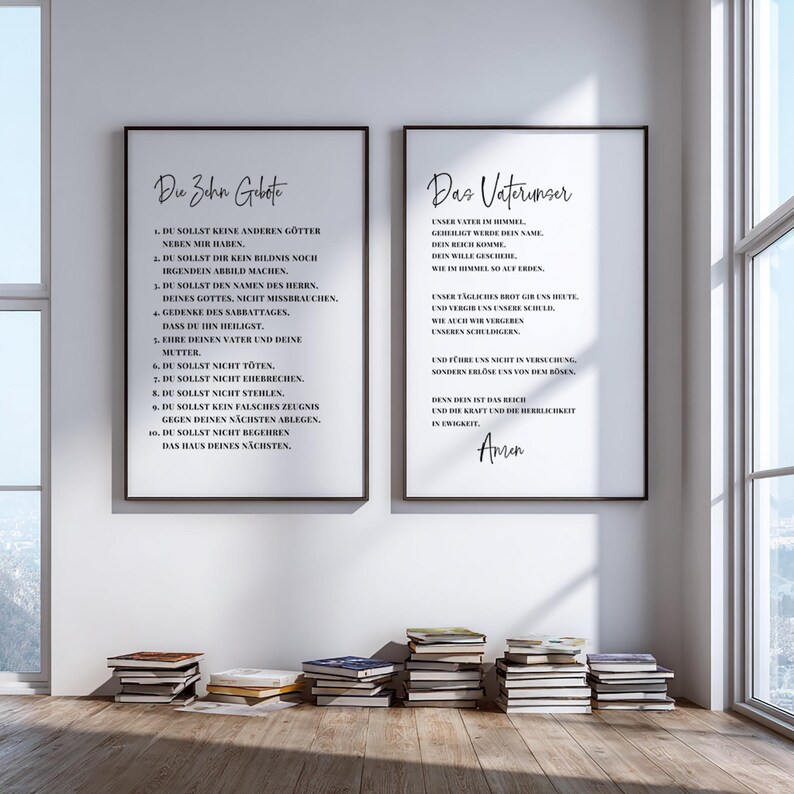 May include: Two framed prints with black frames and white backgrounds hang on a wall. The left print has the text "Die Zehn Gebote" and the right print has the text "Das Vaterunser". Stacks of books are on the floor in front of the wall.