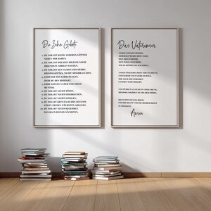 May include: Two framed prints with black text on a white background. The left print says "The Ten Commandments" in German. The right print says "The Lord's Prayer" in German. Several stacks of books are in front of the prints.