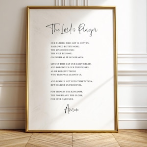 May include: A framed print featuring "The Lord's Prayer" in elegant script. The text is centered on a cream-colored background, enclosed within a gold-colored frame. The print is displayed against a white wall and wooden floor.