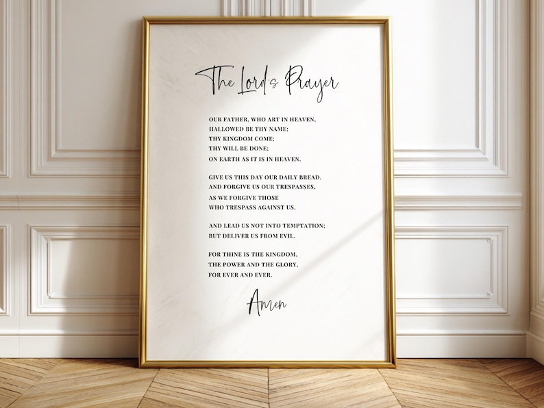 May include: A framed print featuring "The Lord's Prayer" in elegant script. The text is centered on a cream-colored background, enclosed within a gold-colored frame. The print is displayed against a white wall and wooden floor.