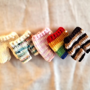 May include: Six miniature knitted shorts in a variety of colours. The shorts include cream, multicoloured, pink, rainbow, and brown and white striped designs. Each pair has a decorative edge.