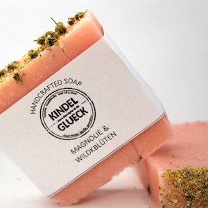 May include: Handmade pink soap bars with dried herbs on top. One bar is angled, displaying a white label with the text "HANDCRAFTED SOAP KINDEL GLUECK MAGNOLIE & WILDKBLÜTEN". The other bar is in the background.