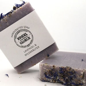 May include: Two bars of lavender and wild herb handcrafted soap. The soap is a light purple color and has dried lavender and herbs embedded in it. The top bar has a white label with the words "Handcrafted Soap" and "Kindel Glueck" in a circle. The label also says "Lavendel & Wildkräuter".
