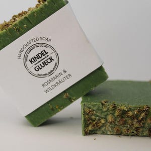 May include: Two bars of green handmade soap with a white label that reads "Handcrafted Soap Kindel Glueck Rosmarin & Wildkraeuter".