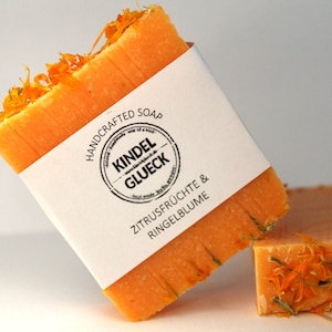 Może przedstawiać: Pomarańczowy ręcznie robiony kawałek mydła z płatkami suszonych kwiatów. Mydło ma białą etykietę z napisem "HANDCRAFTED SOAP KINDEL GLUECK ZITRUSFRÜCHTE & RINGELBLUME". W tle widoczny jest drugi kawałek mydła.