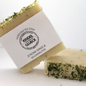 Könnte beinhalten: Zwei handgemachte Seifenstücke mit einem weißen Etikett, auf dem "HANDCRAFTED SOAP" und "KINDEL GLUECK" in einem Kreis mit dem Text "ZITRONE MINZE & WILDKRAUTER" darunter steht.