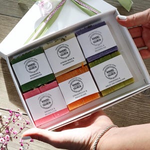 Könnte beinhalten: Eine weiße Geschenkbox mit grünem und rosa Band, gefüllt mit sechs handgefertigten Seifen. Jede Seife trägt das Label "Kindel Glueck" und enthält natürliche Inhaltsstoffe wie Rosmarin, Zitronenminze und Lavendel.