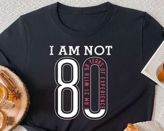 Puede incluir: Una camiseta negra de manga corta con el texto blanco I AM NOT 80 y un número 80 grande. Dentro del cero, un texto rosa dice I AM 21 WITH 59 YEARS OF EXPERIENCE. La camiseta tiene cuello redondo y está hecha de tela de color negro sólido.