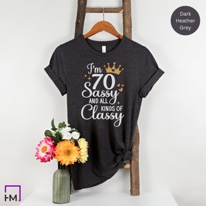 Puede incluir: Camiseta gris oscuro jaspeado con un gr&aacute;fico blanco que dice "I'm 70 Sassy and all kinds of Classy" con una corona dorada y corazones.