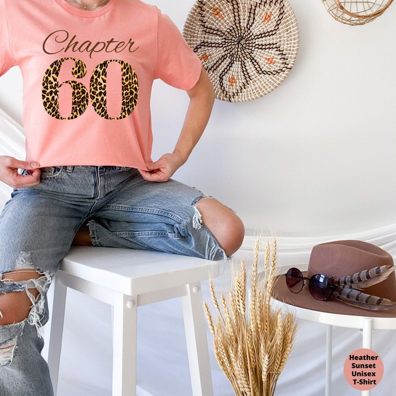 May include: A peach-colored short-sleeve t-shirt featuring the text "Chapter 60" in a dark brown script font above the number "60" in a bold, black and brown leopard print pattern. The shirt is worn with distressed blue denim jeans.