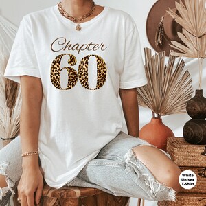 May include: A white short-sleeve t-shirt featuring the brown text "Chapter" above the large number "60" in a leopard print pattern. The shirt is paired with light blue distressed denim jeans, a gold chain necklace, and a gold bracelet.