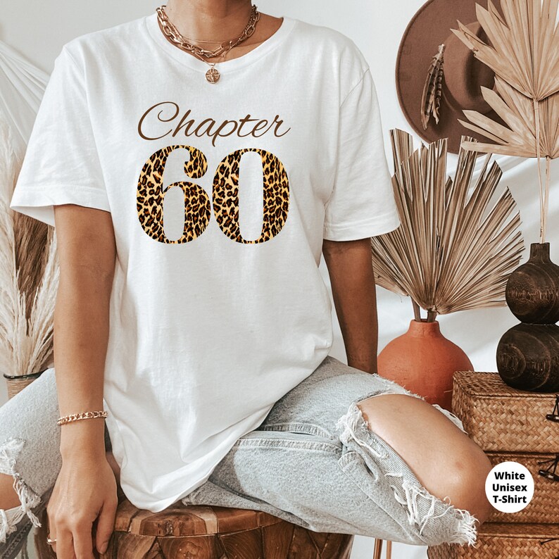 May include: A white short-sleeve t-shirt featuring the brown text "Chapter" above the large number "60" in a leopard print pattern. The shirt is paired with light blue distressed denim jeans, a gold chain necklace, and a gold bracelet.