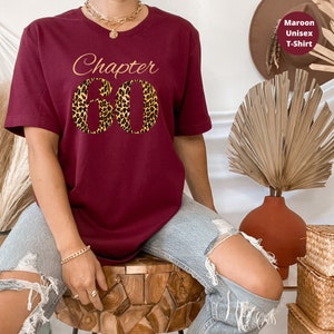 May include: A maroon short-sleeved t-shirt featuring the text "Chapter" in gold cursive script above the number "60" in a bold leopard print pattern. The shirt has a crew neckline and is made of a solid-colored fabric with a smooth, matte finish.