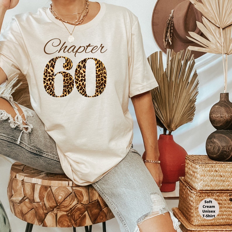 May include: A soft cream-colored unisex t-shirt featuring the text "Chapter 60" in a brown script font, with the number 60 filled in with a brown and black leopard print pattern. The shirt is worn with light blue distressed denim jeans.