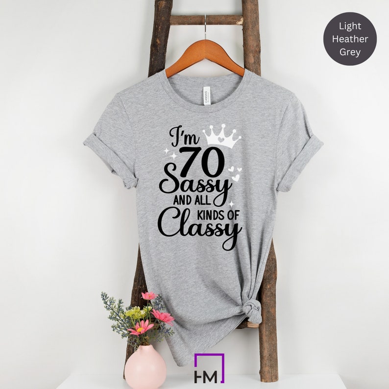 Puede incluir: Una camiseta gris jaspeado claro con un gr&aacute;fico negro que dice "I'm 70 Sassy and all kinds of Classy" con una corona y corazones.