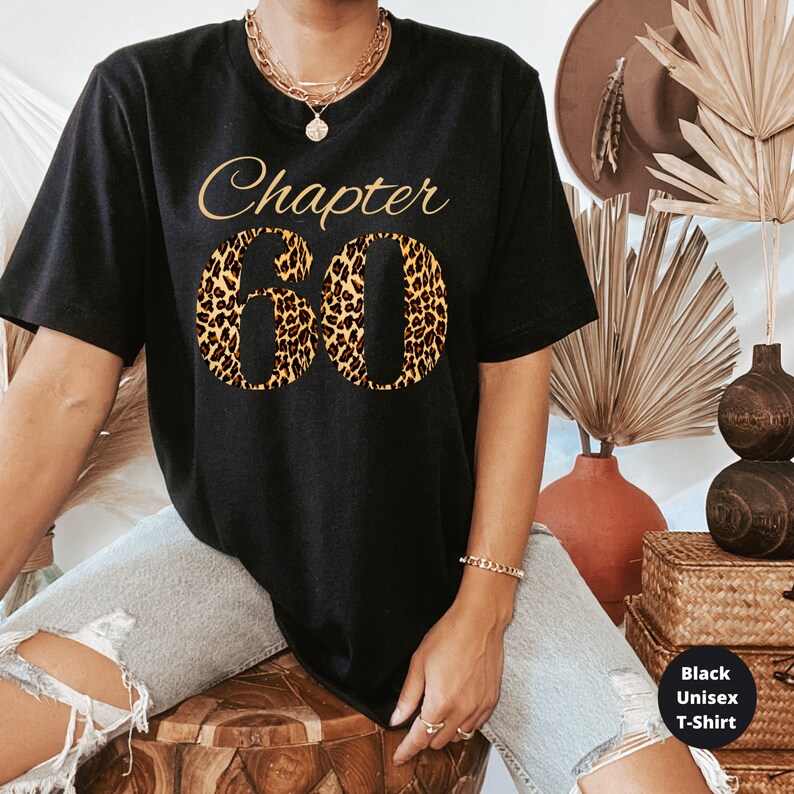 May include: A black short-sleeved t-shirt featuring the text "Chapter" in gold script above the large number "60" filled with a brown and black leopard print pattern. The shirt includes a small black square graphic in the corner with the text "Black Unisex T-Shirt".