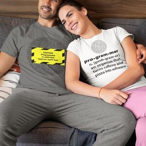 Op de afbeelding: Een stel draagt ​​grijze en witte T-shirts met een humoristische definitie van een programmeur. De tekst op het grijze shirt luidt "LET OP Programmeren is verslavend (en te veel plezier)". De tekst op het witte shirt luidt "pro-gram.mer n. [proh-gram-er] een organisme dat cafeïne en pizza omzet in software".