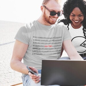 May include: Two people are sitting on a bench, looking at a laptop. The person in the front is wearing a grey t-shirt with a binary code design that includes the word "HACKED" in red. The person in the back is wearing a white t-shirt with a black circle design.