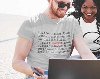 Pode incluir: Duas pessoas estão sentadas em um banco, olhando para um laptop. A pessoa na frente está vestindo uma camiseta cinza com um design de código binário que inclui a palavra "HACKED" em vermelho. A pessoa atrás está vestindo uma camiseta branca com um design de círculo preto.
