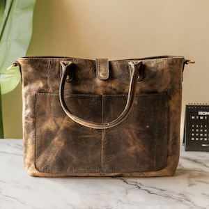 May include: A brown leather tote bag with two pockets on the front and a top handle. The bag has a distressed look and is made of a thick, durable leather.