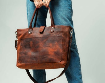 Könnte beinhalten: Eine braune Ledertasche mit langem Trageriemen. Die Tasche hat einen Klappverschluss und eine Fronttasche. Die Tasche wird von einer Person getragen, die blaue Jeans trägt.