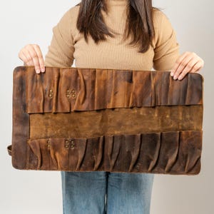 May include: A brown leather tool roll with multiple pockets and brass rivets. The roll is held open, displaying the interior pockets designed to hold various tools. The leather has a distressed appearance.