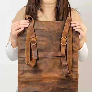 May include: A brown leather roll-up bag with adjustable straps and brass-colored buckles. The bag is held open, showcasing its interior. The leather has a distressed finish, giving it a vintage look. The bag appears to be designed for carrying tools or other items.