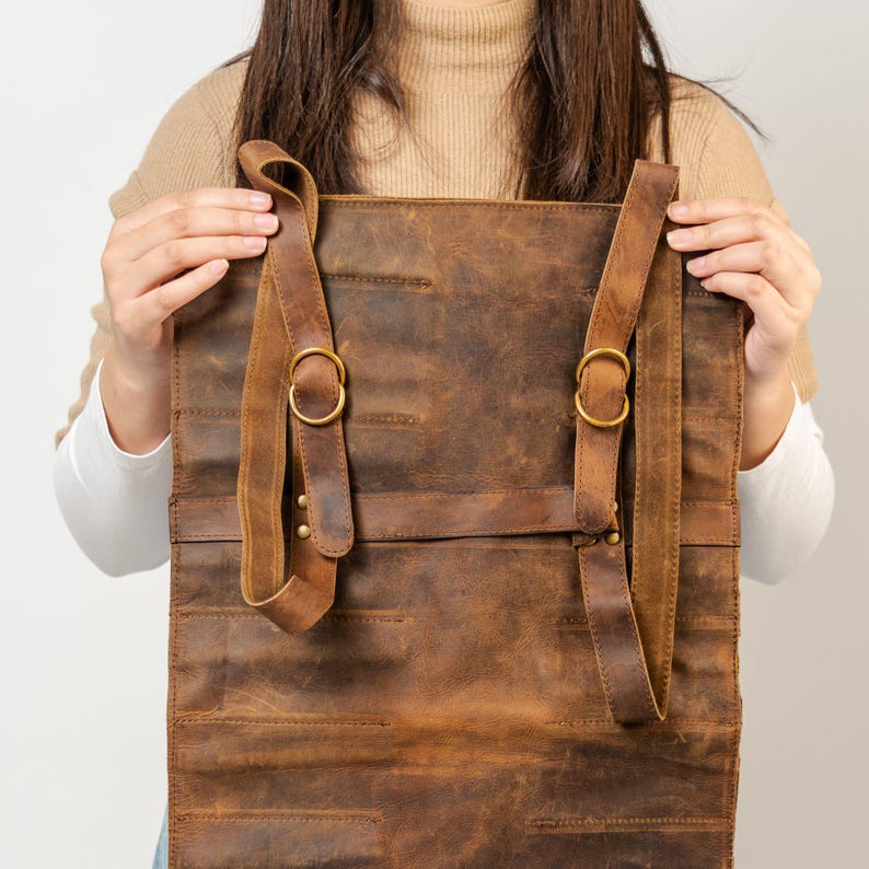 May include: A brown leather roll-up bag with adjustable straps and brass-colored buckles. The bag is held open, showcasing its interior. The leather has a distressed finish, giving it a vintage look. The bag appears to be designed for carrying tools or other items.
