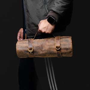 May include: A brown leather roll bag with two leather straps and gold-colored buckles. The bag is cylindrical and has a distressed finish. It is being held by a person wearing a dark jacket and a smartwatch.