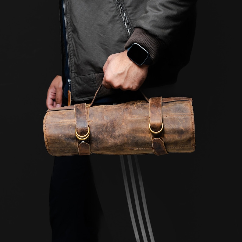 May include: A brown leather roll bag with two leather straps and gold-colored buckles. The bag is cylindrical and has a distressed finish. It is being held by a person wearing a dark jacket and a smartwatch.