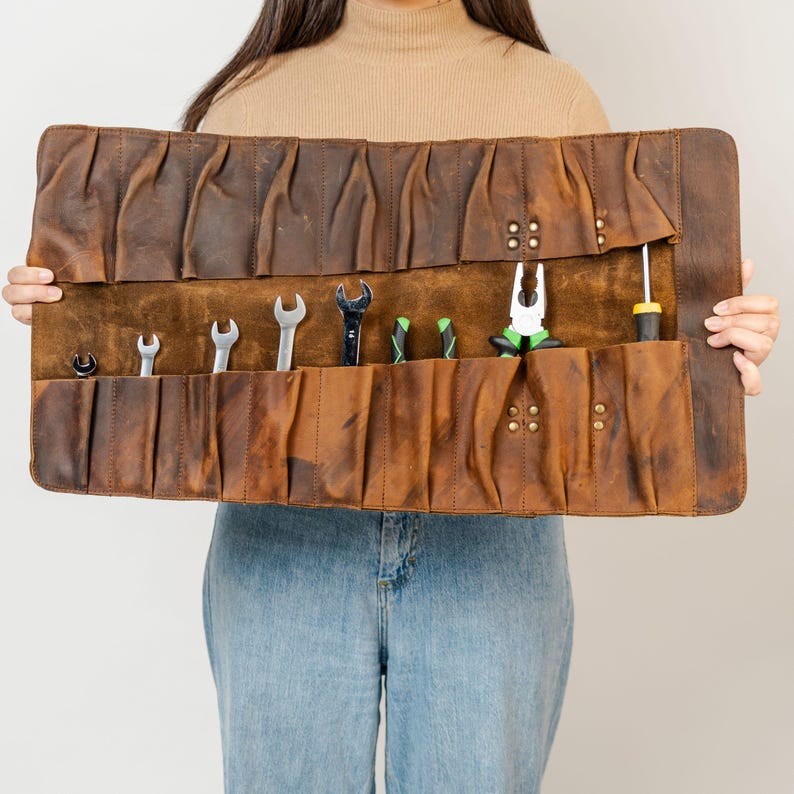 May include: A brown leather tool roll with various tools, including wrenches, pliers, and a screwdriver. The roll is held open, displaying the tools in individual pockets. The leather has a distressed finish.