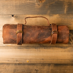May include: A rolled-up, brown leather knife roll with a handle and two straps secured with brass-colored buckles. The leather has a distressed appearance, and the roll is lying on a wooden surface.
