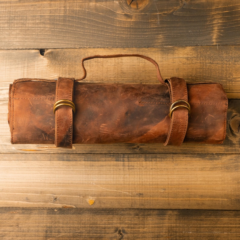 May include: A rolled-up, brown leather knife roll with a handle and two straps secured with brass-colored buckles. The leather has a distressed appearance, and the roll is lying on a wooden surface.