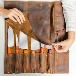 May include: A brown leather knife roll containing six knives. The knives have silver blades and orange and brown handles. The roll is open, showing the knives, and has a zip closure.