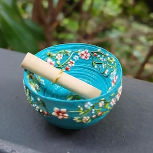 May include: A small, turquoise bowl with a floral design and a rolled-up paper scroll inside. The bowl is decorated with white, red, and pink flowers and green leaves. A gold ring encircles the scroll.