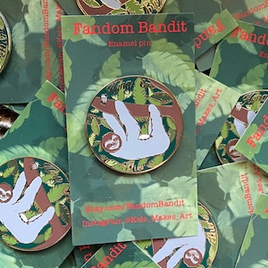 May include: A circular enamel pin depicting a light grey sloth on a brown branch, encircled by green leaves. The pin has a gold edge and the words "Hang Cher". It rests on a green card with "Fandom Bandit" and social media details.