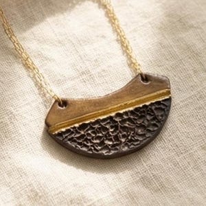 Puede incluir: Un collar dorado con un colgante en forma de media luna. El colgante tiene una sección inferior texturizada, de color marrón oscuro, y una sección superior lisa, dorada. El collar se muestra sobre una tela color crema.