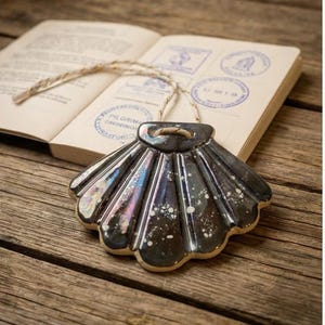 Puede incluir: Un adorno decorativo de concha iridiscente con un cordón, sobre una superficie de madera. La concha es oscura con motas blancas y un brillo nacarado. Un libro abierto con sellos está en el fondo.