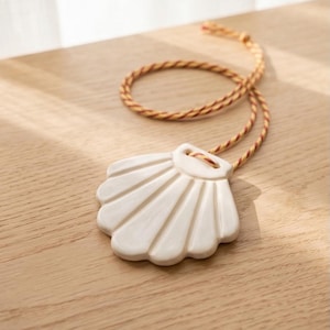 Puede incluir: Un adorno de concha de cerámica blanca con un diseño acanalado, suspendido por un cordón trenzado en tonos amarillos y rojos. El adorno descansa sobre una superficie de madera clara, con sombras de luz natural.