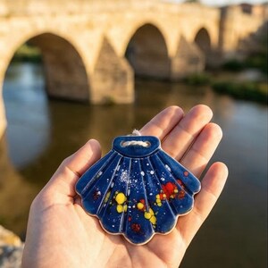 Puede incluir: Un colgante de cerámica azul con forma de concha de vieira con salpicaduras de pintura amarilla, roja y blanca. La concha se sostiene en una mano, con un puente de piedra y un río al fondo. El colgante tiene un pequeño agujero para una cuerda.