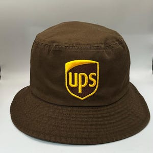 May include: A brown bucket hat featuring a centered, embroidered UPS logo patch. The logo consists of a yellow shield outline containing the lowercase letters ups in yellow, set against a dark brown background within the shield shape.