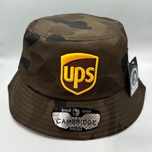 May include: Camouflage bucket hat with the UPS logo patch. The hat is brown and black with a camouflage pattern. The UPS logo is yellow and brown. The hat has a tag that says "CAMBRIDGE" and "PB261".