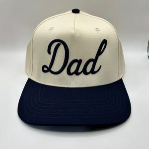 May include: A cream-colored baseball cap with a flat navy blue brim and a small navy blue button on top. The front panel features the word Dad embroidered in a dark navy blue cursive script font, centered prominently on the light fabric surface.