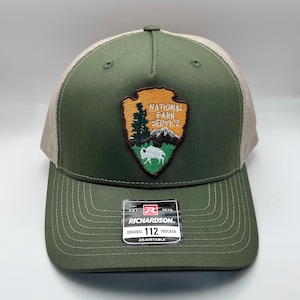 May include: Green and tan trucker hat with the National Park Service logo. The hat features a green front panel and brim, a tan mesh back, and an adjustable strap. The logo is an arrowhead shape with a tree, mountains, and a bison.
